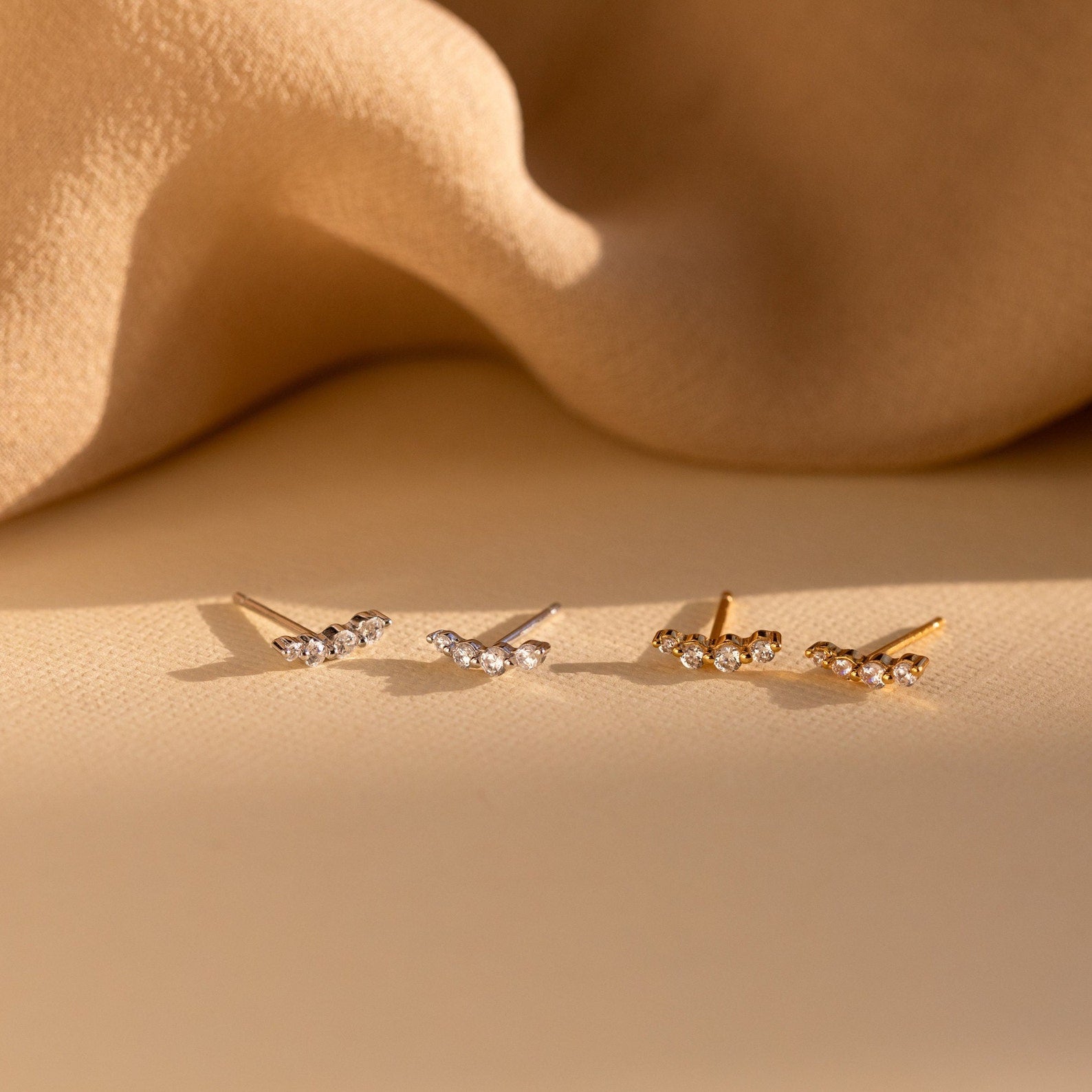 Two pairs of elegant Tiny Diamond Climbers earrings—one in silver and one in gold—feature delicate crystals on a beige fabric background, making them a perfect anniversary gift.