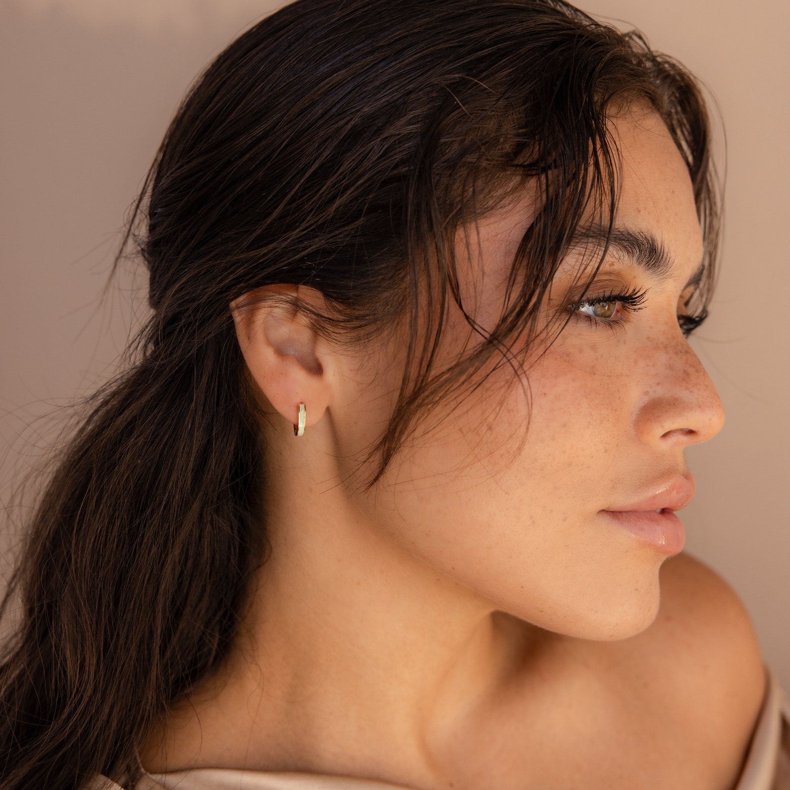 A woman with long dark hair and freckles wears shimmering Opal Inlay Earrings, gazing to the side against a beige background.