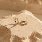 Two small gold hoop earrings with embedded stones rest on a textured beige stone, highlighting the radiant charm of Citrine Pave Huggies.