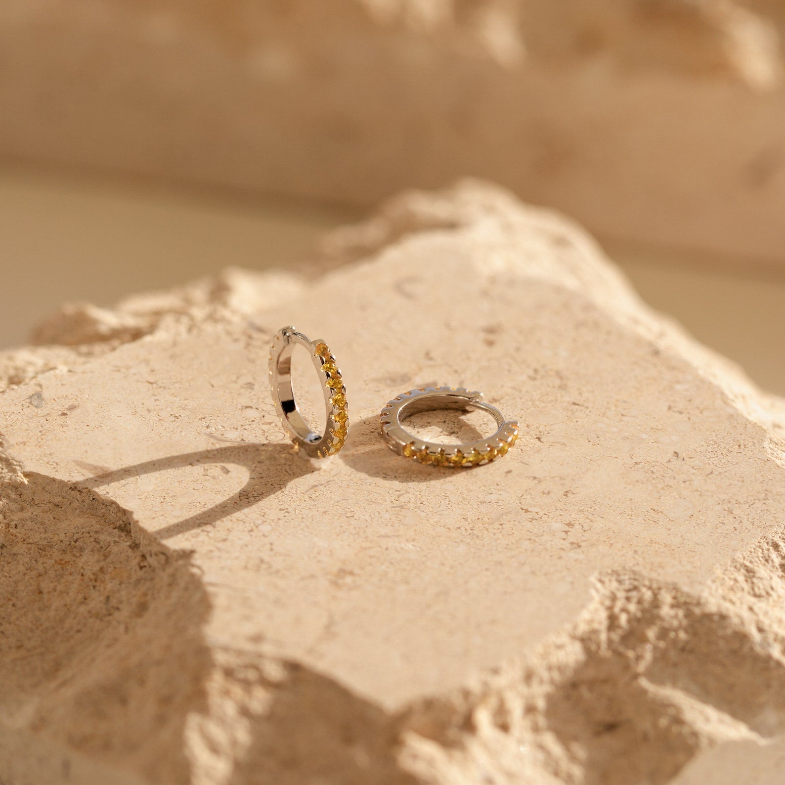 Two small gold hoop earrings with embedded stones rest on a textured beige stone, highlighting the radiant charm of Citrine Pave Huggies.