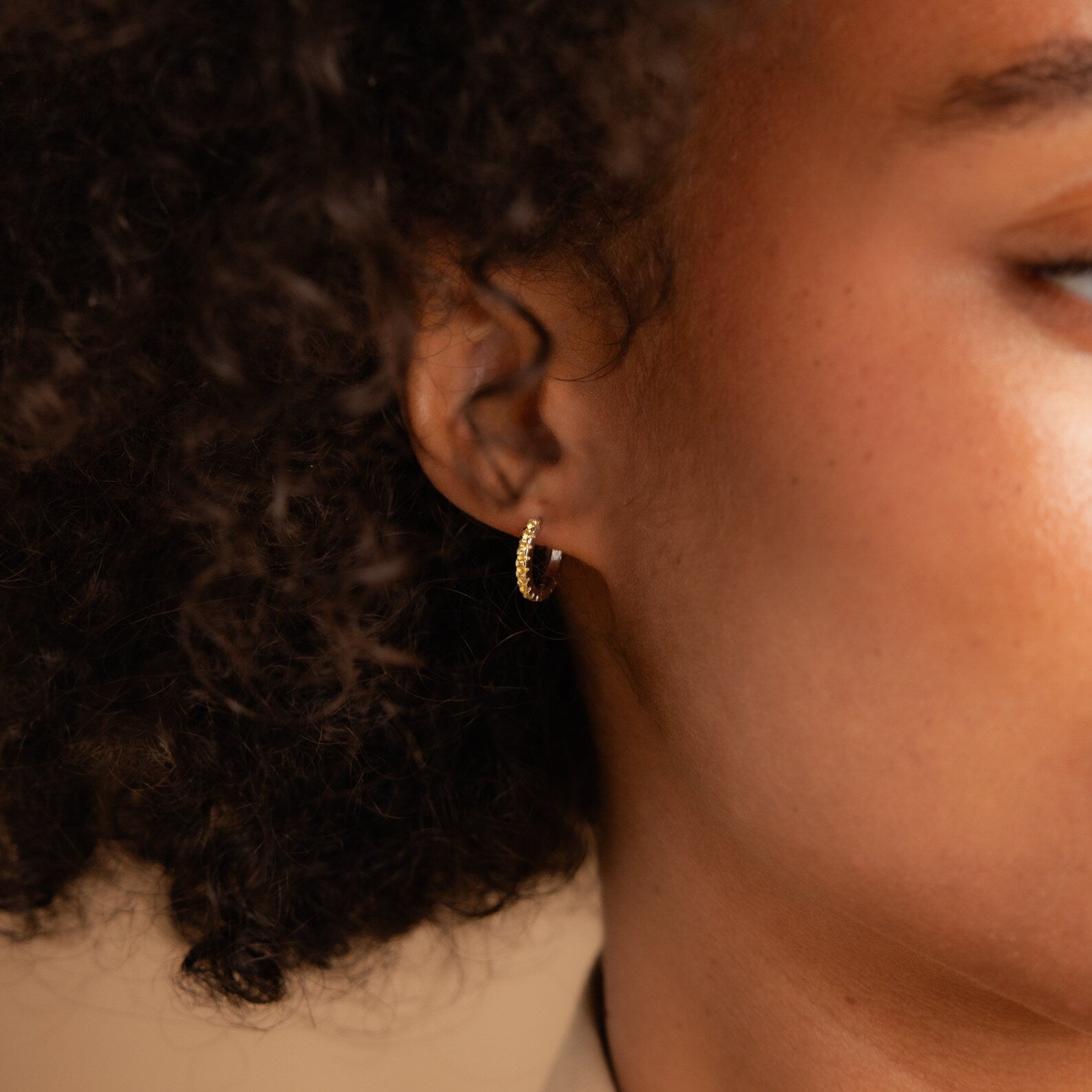Close-up of a person with curly hair wearing Citrine Pave Huggies in their left ear.