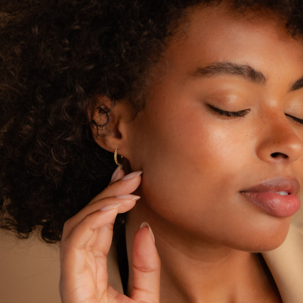 A woman with curly hair and closed eyes touches her face, showcasing her Citrine Pave Huggies—delicate earrings that bring a subtle sparkle to her look.