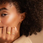 A woman with curly hair wears gold rings and small hoop earrings, resting her chin on her hand. Her look is brightened by the Citrine Pave Huggies.