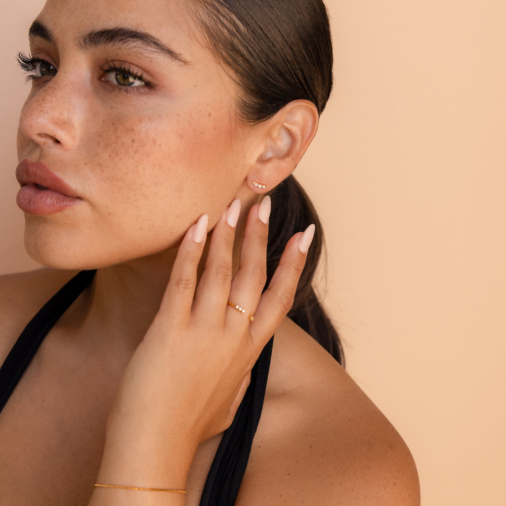 Woman with slicked-back hair, wearing elegant earrings and gold jewelry, touches her face against a beige background—making Tiny Diamond Climbers the perfect anniversary gift.
