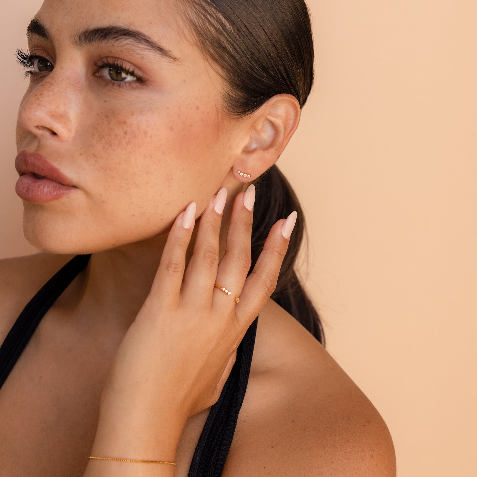 Woman with slicked-back hair, wearing elegant earrings and gold jewelry, touches her face against a beige background—making Tiny Diamond Climbers the perfect anniversary gift.
