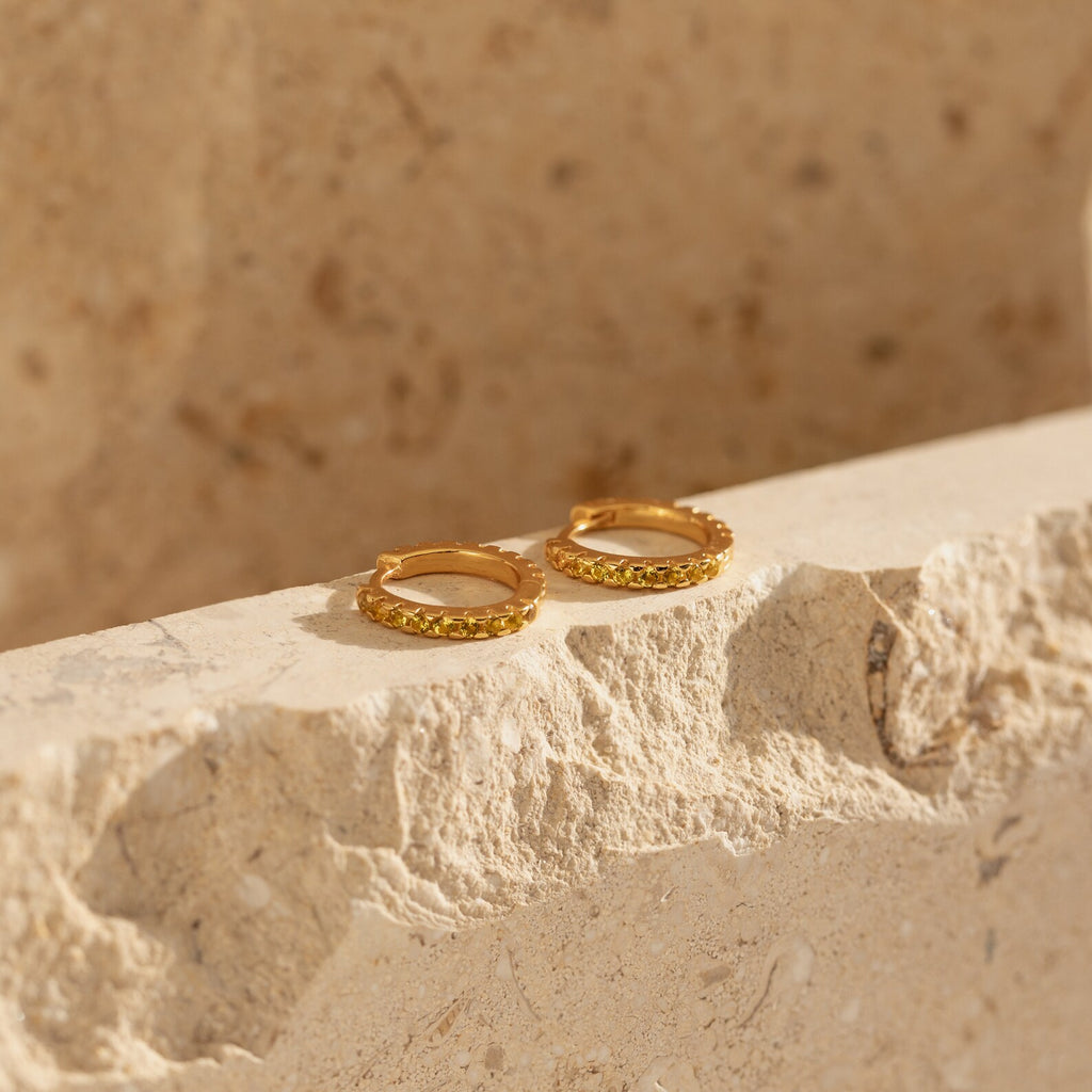 The Citrine Pave Huggies are displayed on a textured beige stone surface, their elegant hoops glowing in natural sunlight.