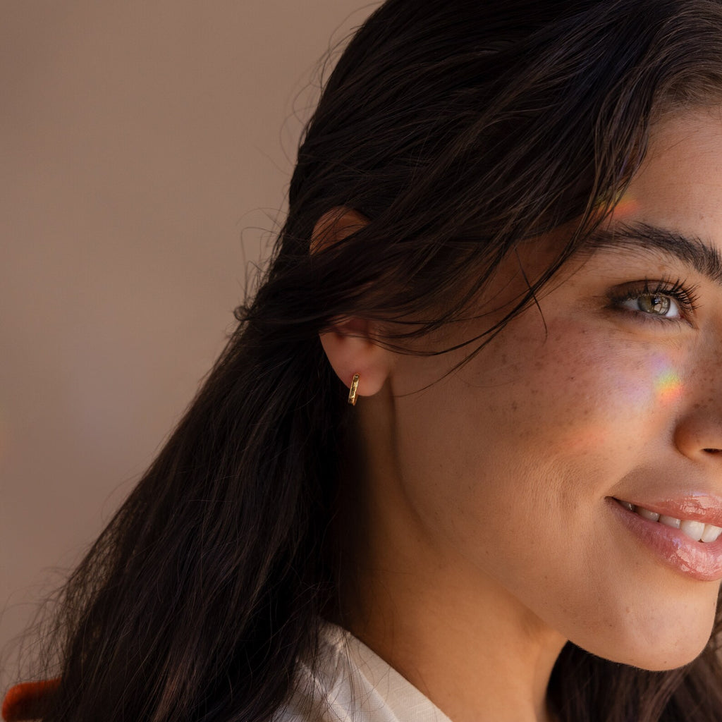 Smiling woman with dark hair wears Crescent Moon Opal Huggies, a small hoop earring, as a rainbow light shines on her cheek.