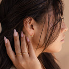 A woman with long dark hair touches her ear, wearing Crescent Moon Opal Huggies and gold rings, paired with a nude manicure.