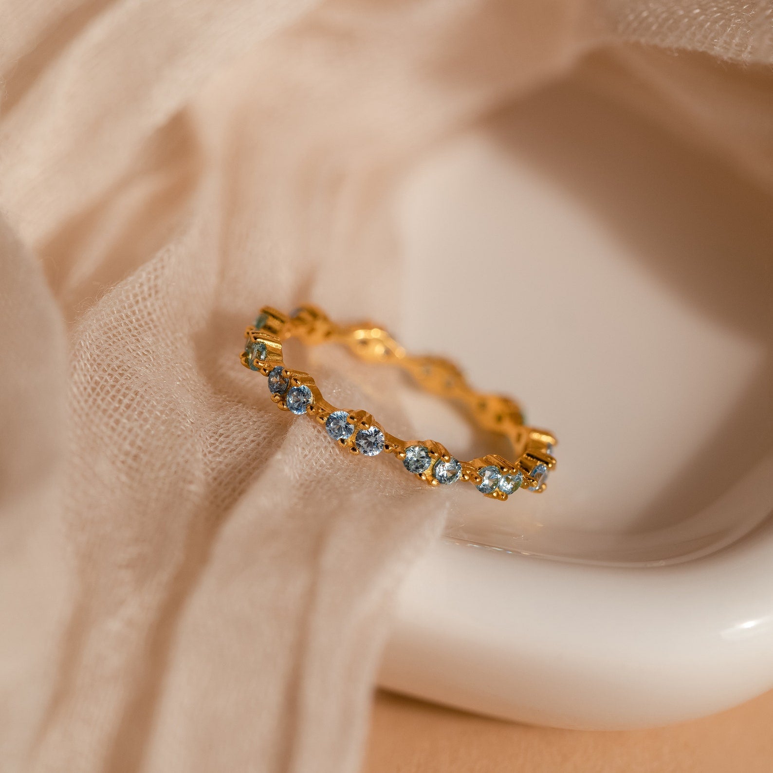 The Aquamarine Eternity Ring glows with blue gemstones, placed on soft beige fabric and a white ceramic dish—an elegant stacking ring choice.
