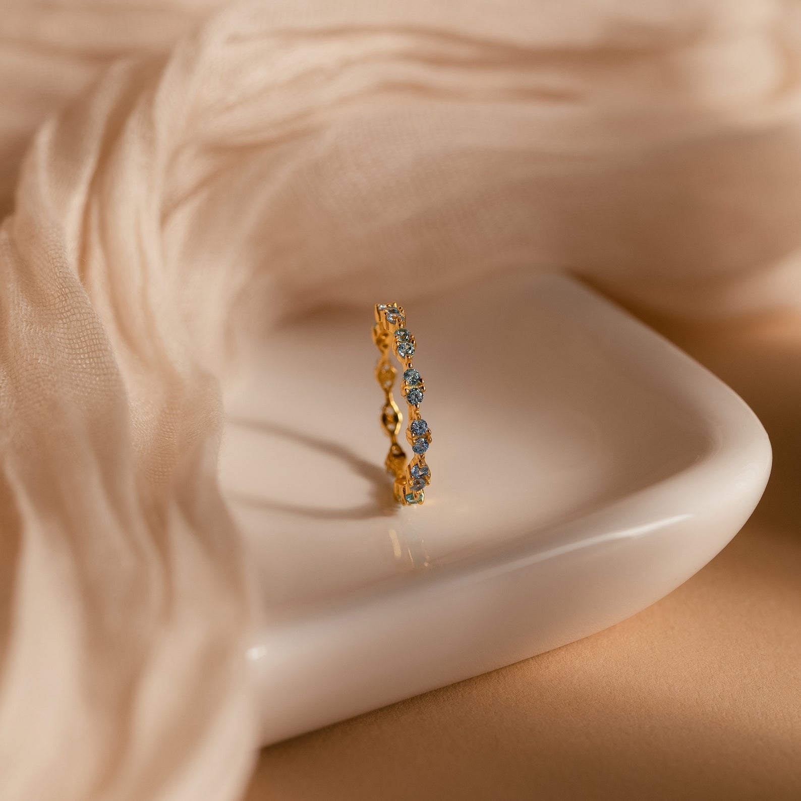 Aquamarine Eternity Ring with sparkling blue gemstones, shown on a white dish with a beige fabric backdrop—an elegant choice for everyday wear.