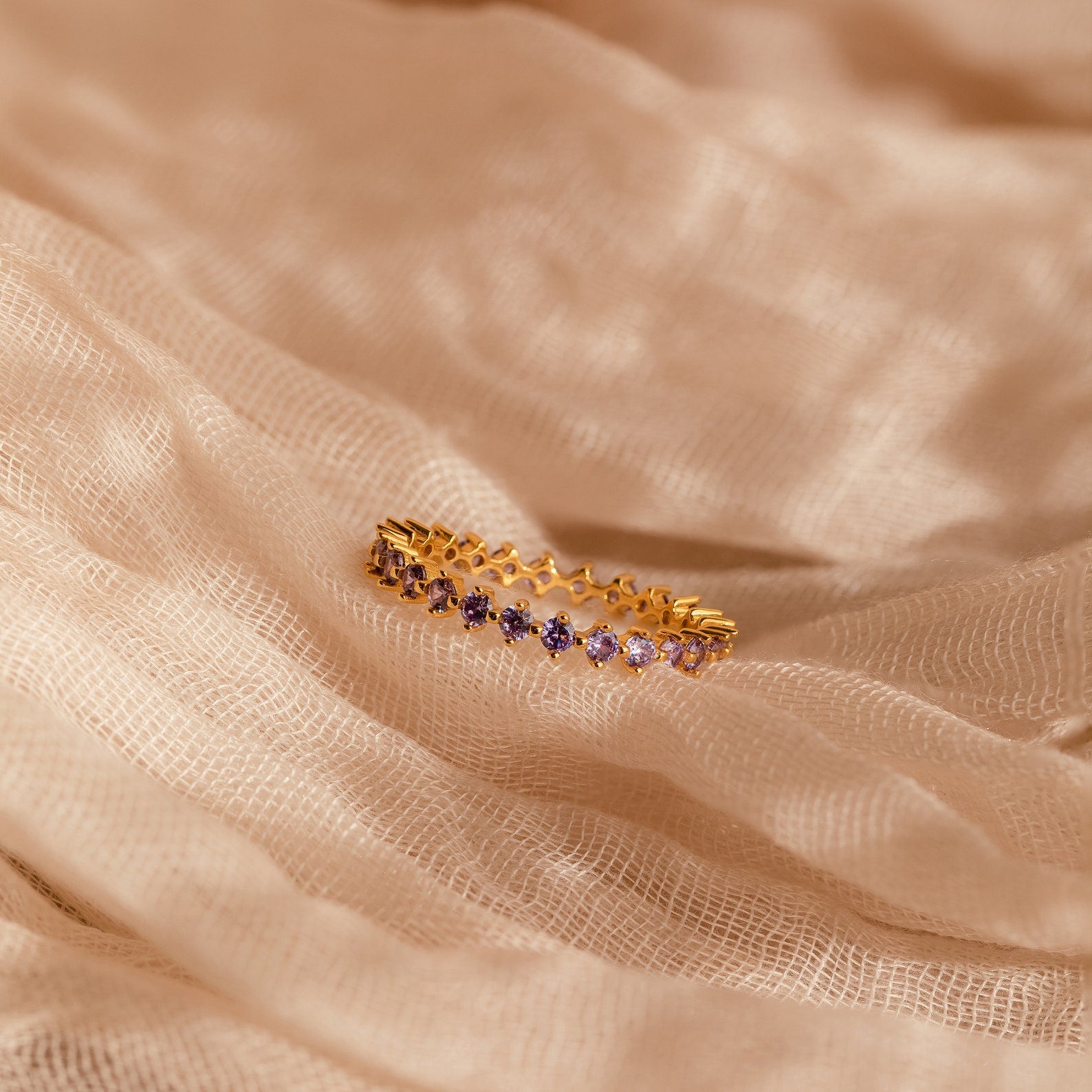 The Amethyst Eternity Ring, a minimalist piece with purple amethyst gemstones, is elegantly showcased on soft beige textured fabric.
