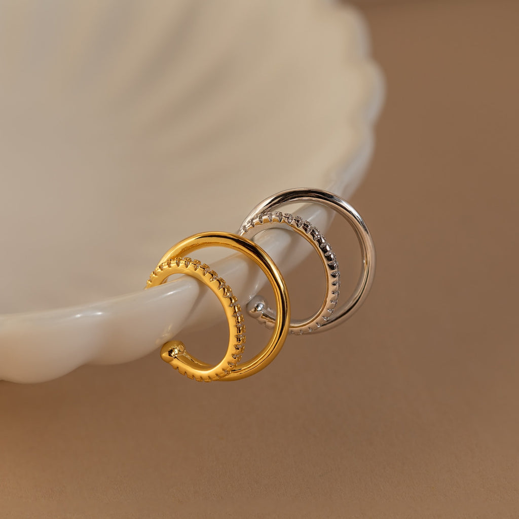 The Lydia Duo Cuffs, featuring gold and silver textured hoops, are displayed on a cream-colored dish beside multilayered earrings for a modern touch.