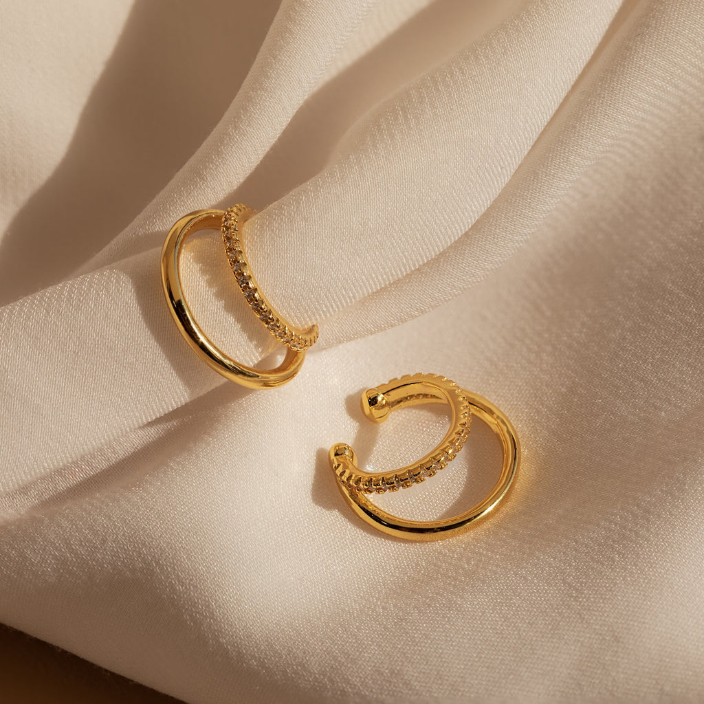 Two gold hoop earrings with small crystals rest on soft, cream-colored fabric, capturing the elegance of Lydia Duo Cuffs.