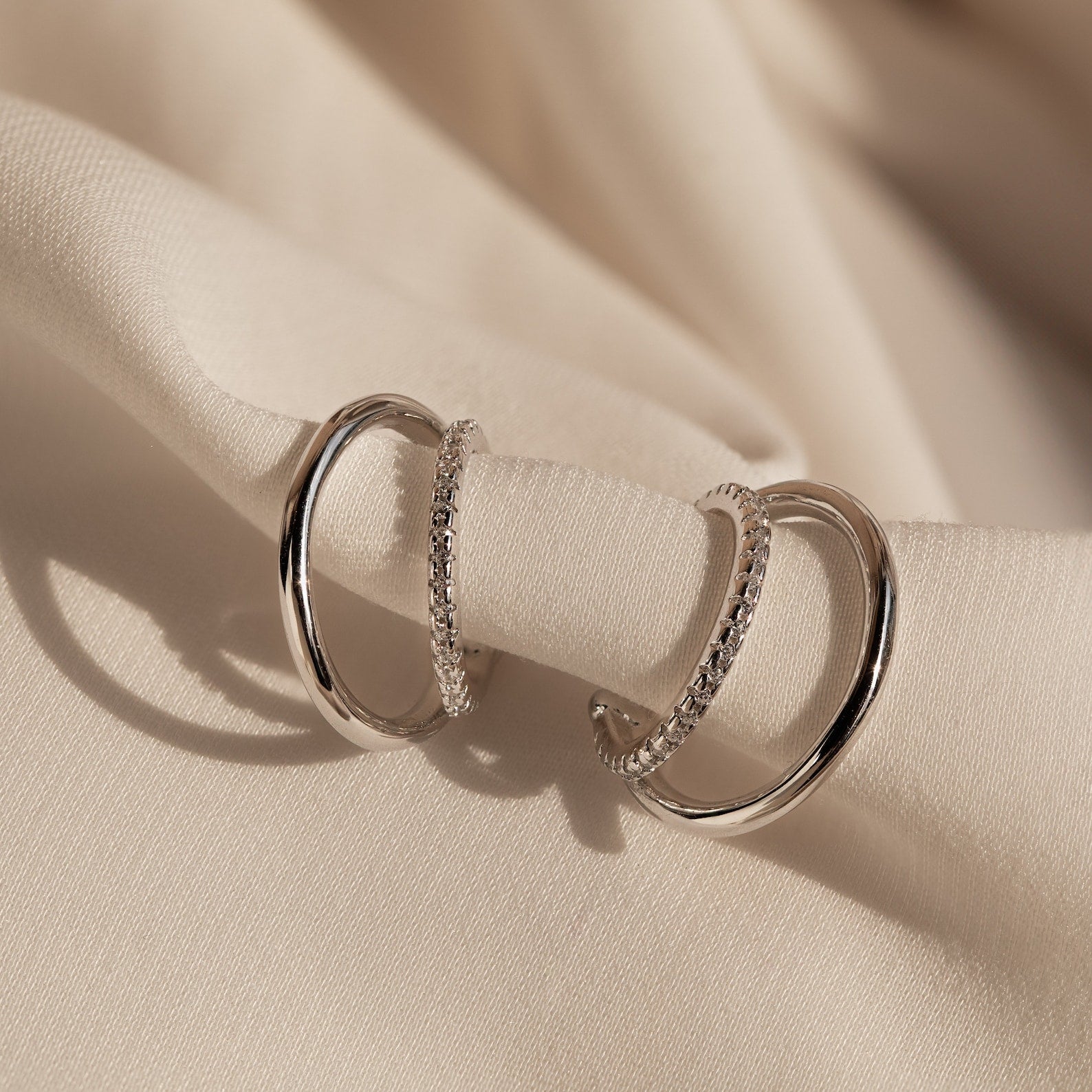 The Lydia Duo Cuffs—two silver hoop earrings with textured details—are elegantly displayed on soft beige fabric, perfect for pairing with multilayered earrings.