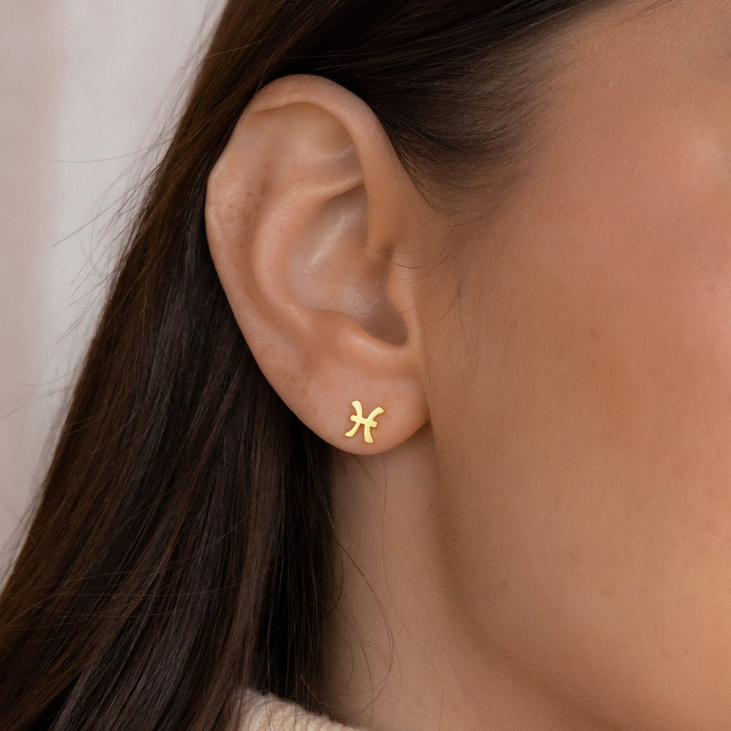 Close-up of a woman's ear wearing a gold Pisces Tiny Zodiac Stud, perfect for your personalized earring stack or pairing with other Tiny Zodiac Studs for a unique star sign look.