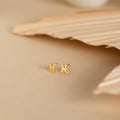 Tiny Zodiac Studs: small gold Pisces star sign earrings on a beige surface with textured leaf decoration in the background—perfect for adding a personalized touch to your earring stack.