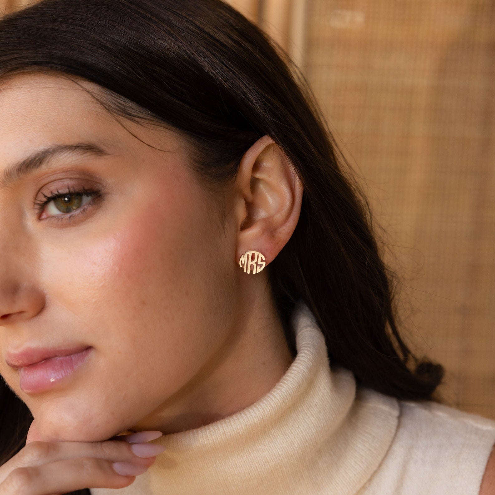 A woman wearing a cream turtleneck and Block Monogram Earrings in a gold geometric design shaped like the word MRS, posing thoughtfully.