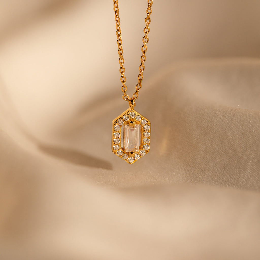 The Daydreamer Diamond Necklace features a gold pendant with a rectangular center stone framed by sparkling diamonds, exuding timeless elegance on a soft beige fabric background.