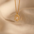 The Daydreamer Diamond Necklace features a gold pendant with a rectangular center stone framed by sparkling diamonds, exuding timeless elegance on a soft beige fabric background.