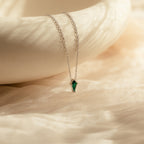 The Art Deco Pendant Necklace features a delicate silver chain and a small green gemstone pendant, elegantly displayed on soft, beige textured fabric.