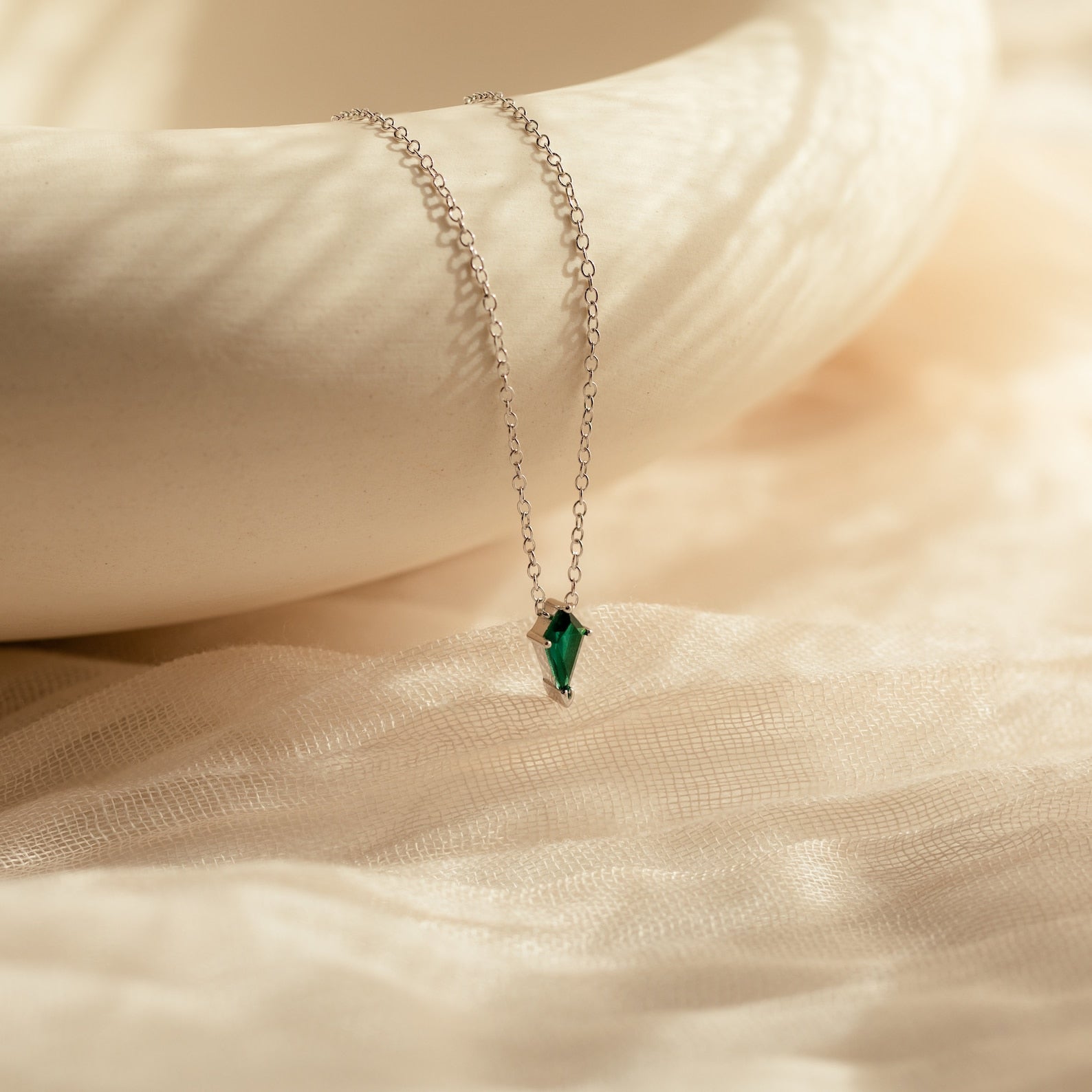 The Art Deco Pendant Necklace features a delicate silver chain and a small green gemstone pendant, elegantly displayed on soft, beige textured fabric.