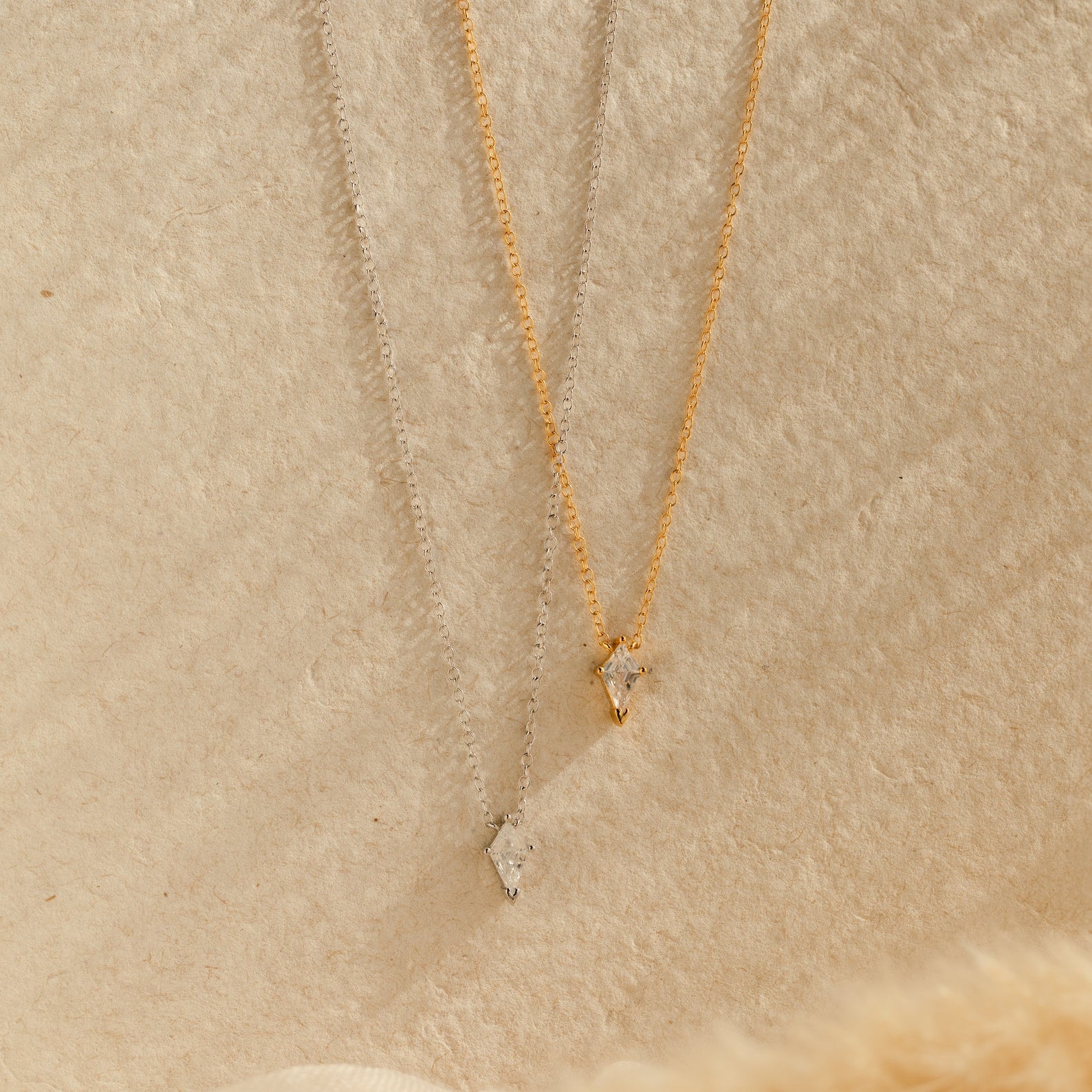 Two Art Deco Pendant Necklaces, one in silver and one in gold, each with a small solitaire pendant, displayed on a textured beige surface.