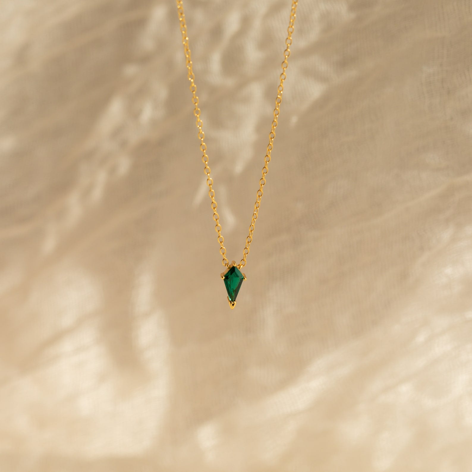 The Art Deco Pendant Necklace, featuring a small green gemstone on a delicate gold chain, rests against a soft beige background.