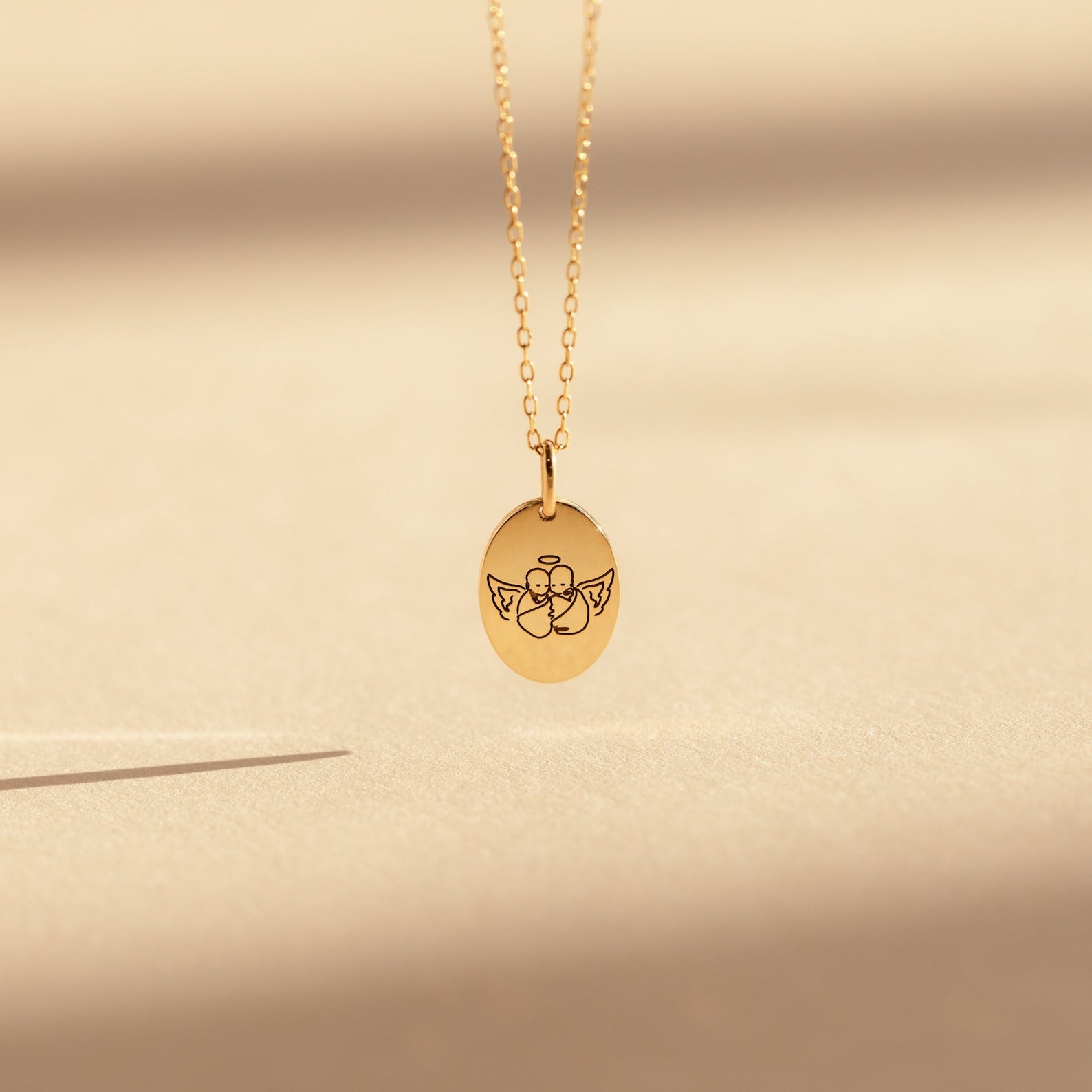 The Child Portrait Necklace is a gold engraved oval pendant with a simple angel design, perfect as a memorial or keepsake piece. Shown against a soft beige background.