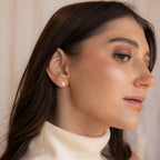 Woman with long brown hair in a cream turtleneck wears Tiny Zodiac Studs—gold Pisces star sign earrings—perfect for a personalized earring stack, shown in side profile.