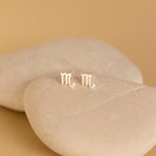 Tiny Zodiac Studs, perfect for personalized earring stacks, displayed on smooth beige stones against a neutral background.