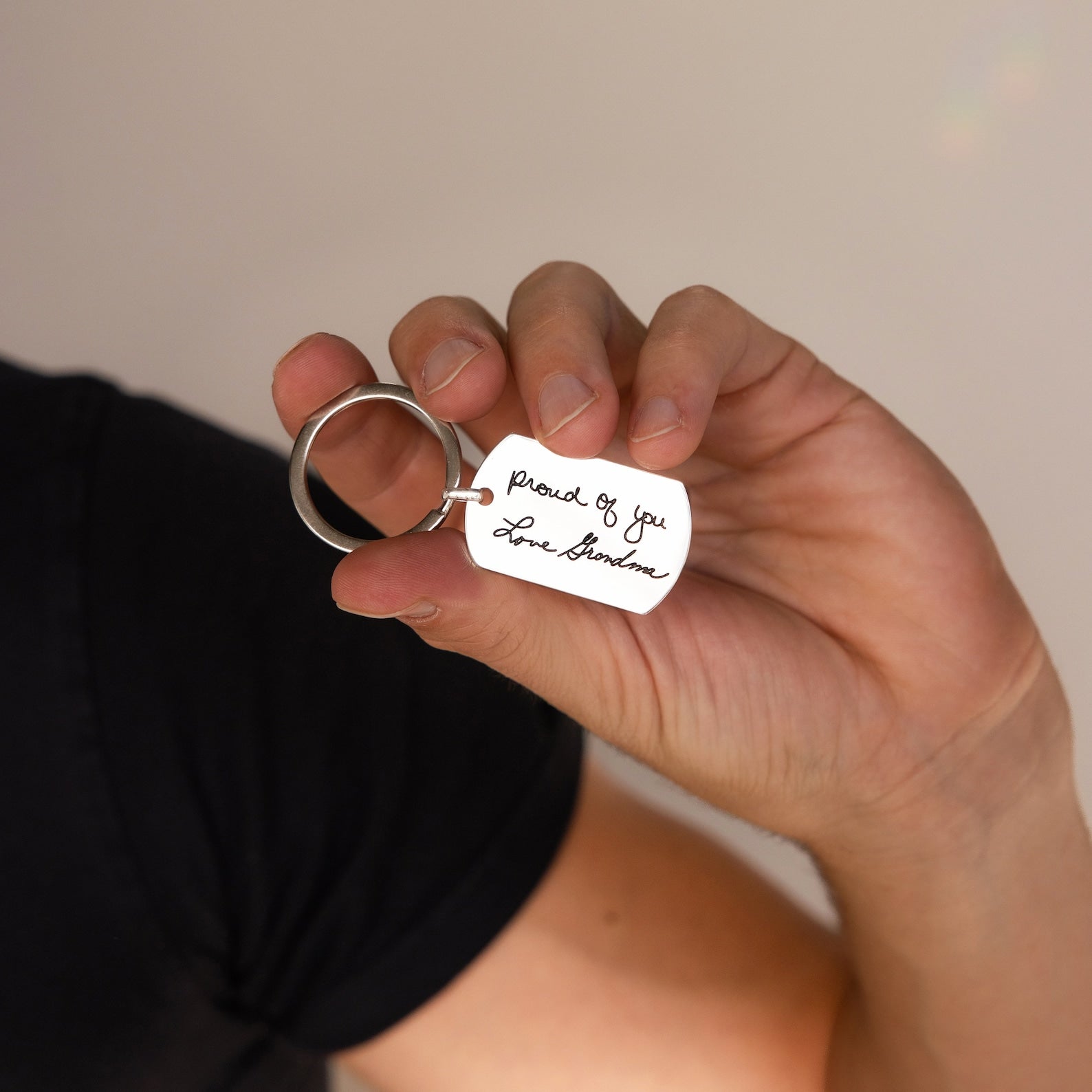 A hand holds a Handwriting Dog Tag Keychain engraved with Proud of you. Love, Grandma in cursive—a personalized and heartfelt Father’s Day gift.