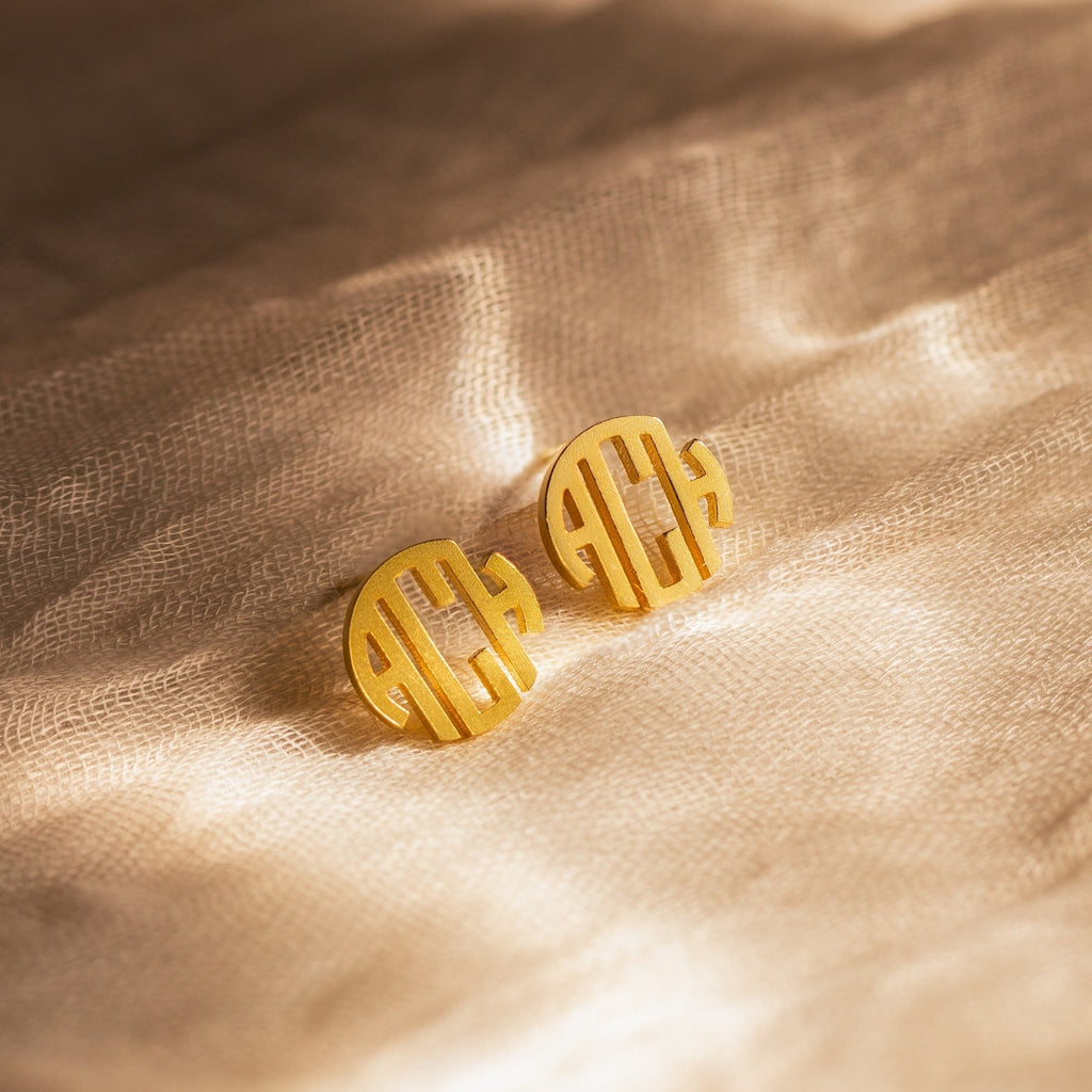 Block Monogram Earrings feature gold round shapes with cut-out initials, adding a geometric touch and shown on textured beige fabric.