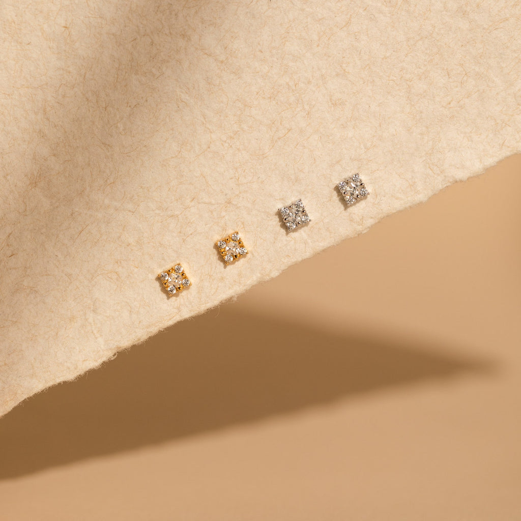 Four pairs of Daydreamer Diamond Studs—two gold and two silver square styles—are displayed on a textured beige surface, adding a dainty, glamorous touch to any look.