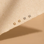 Four pairs of Daydreamer Diamond Studs—two gold and two silver square styles—are displayed on a textured beige surface, adding a dainty, glamorous touch to any look.