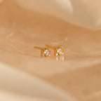 The Daydreamer Diamond Studs, featuring sparkling round gemstones in a glamorous gold setting, rest elegantly on a soft beige surface.