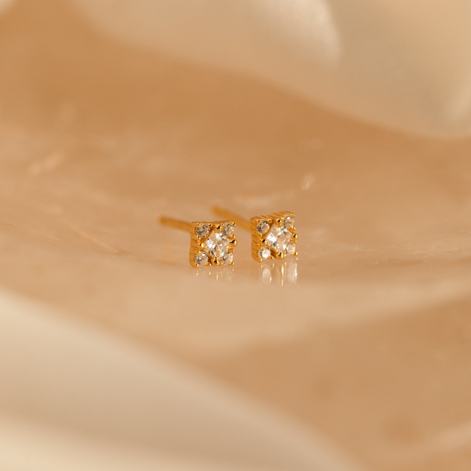 The Daydreamer Diamond Studs, featuring sparkling round gemstones in a glamorous gold setting, rest elegantly on a soft beige surface.