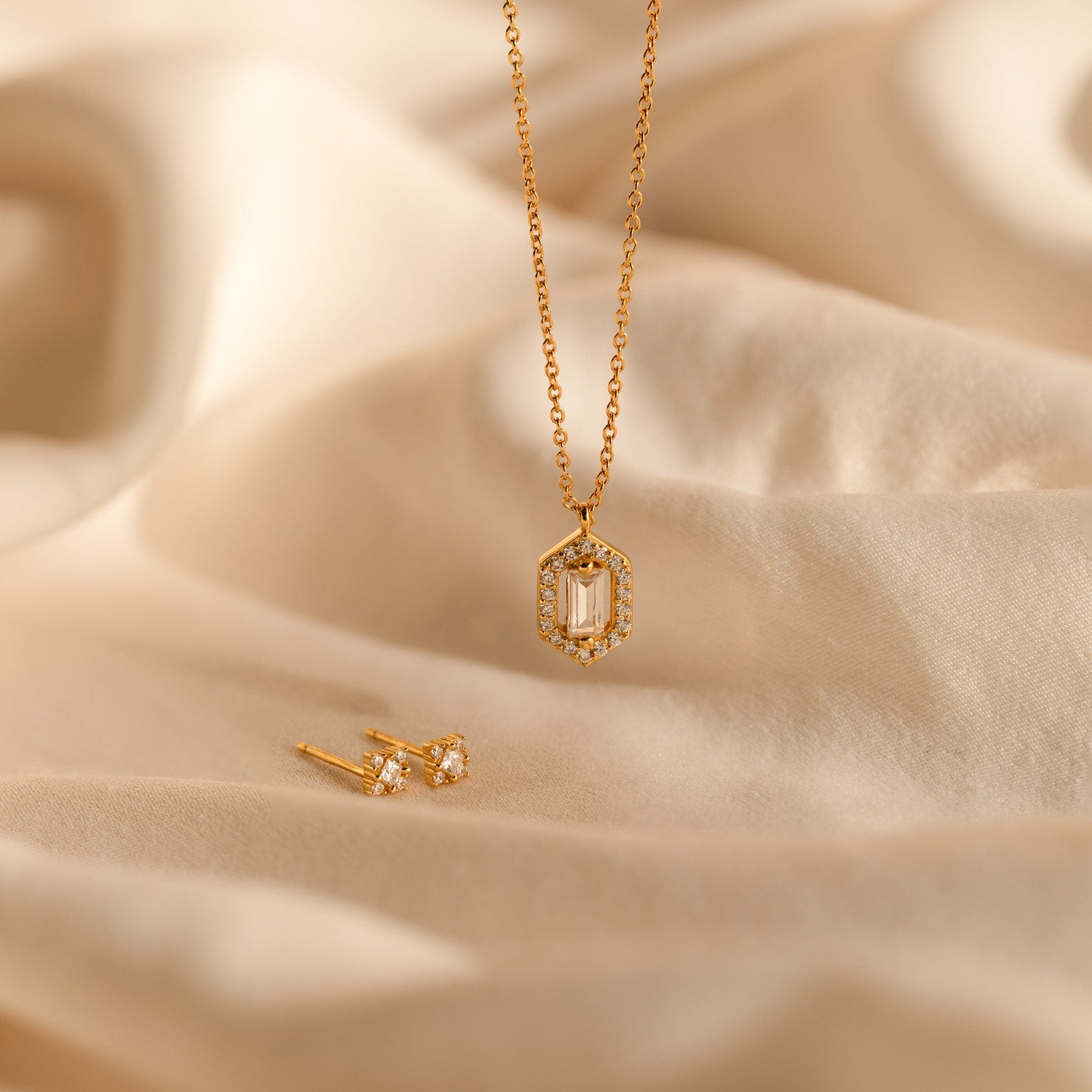 The Daydreamer Diamond Necklace features a hexagon pendant and matching stud earrings, displayed on soft beige fabric for a timelessly elegant and luxurious jewelry set.