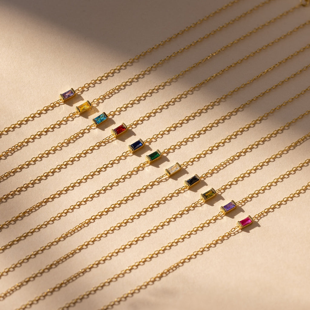 Nine Baguette Birthstone Bracelets featuring colorful rectangular gemstone charms, including personalized options, are arranged in parallel rows on a beige surface.