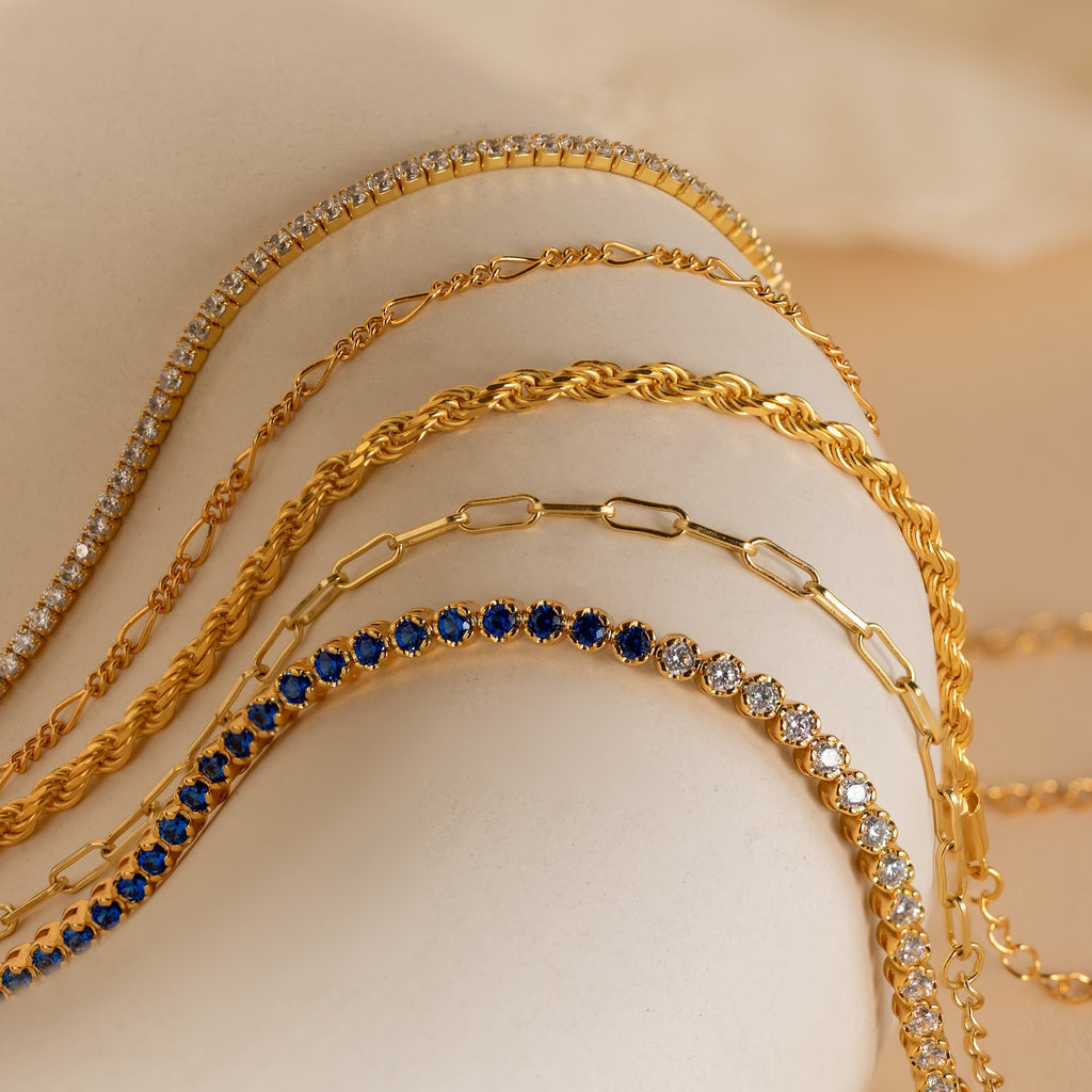 Five gold and gemstone necklaces are displayed on a beige surface in soft lighting, accompanied by Must-Have Bracelet Chains that add an elegant touch of sparkle.