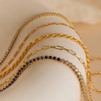 Five gold and gemstone necklaces are displayed on a beige surface in soft lighting, accompanied by Must-Have Bracelet Chains that add an elegant touch of sparkle.