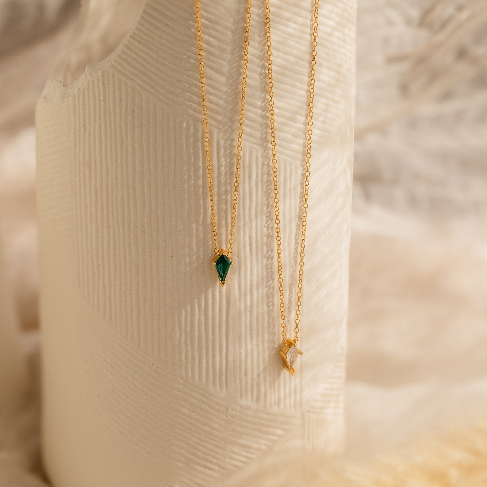 Gold Art Deco pendant necklaces with delicate chains, featuring one green gemstone pendant and one clear gemstone pendant, displayed against a textured ivory backdrop.