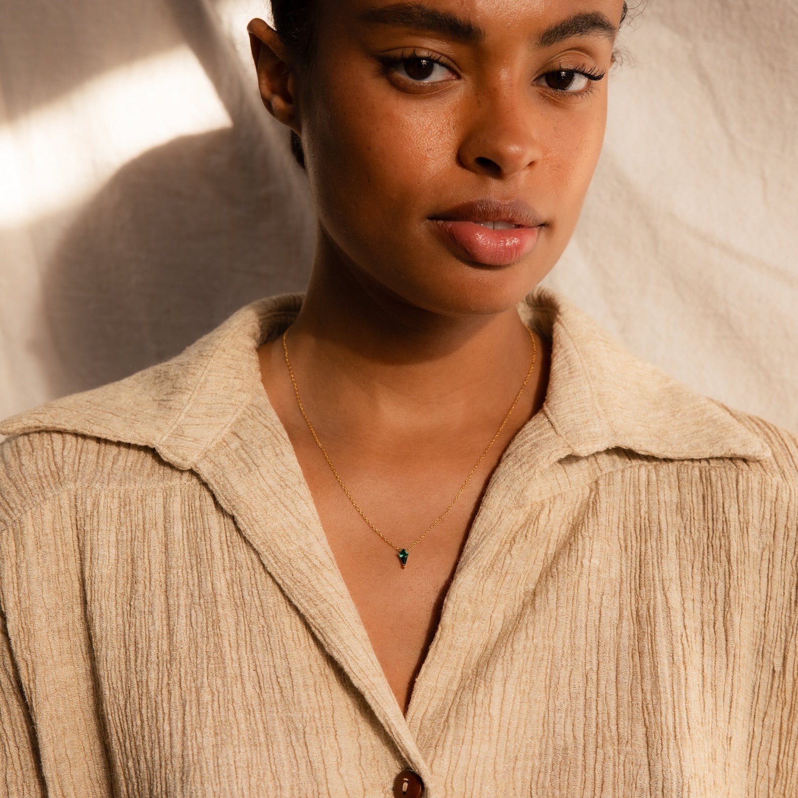 A woman indoors wears a beige textured shirt and the Art Deco Pendant Necklace featuring a small green pendant.