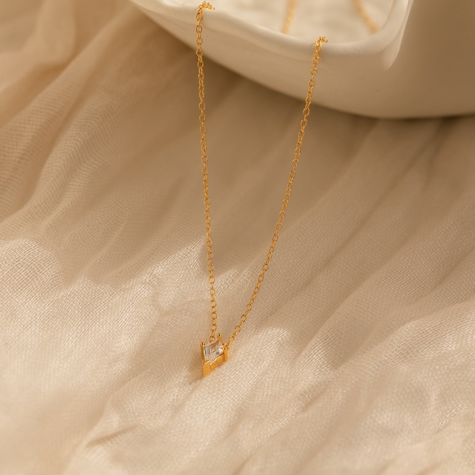 The Art Deco Pendant Necklace features a gold chain and a small geometric kite pendant, shown on soft beige fabric beside a white dish.