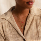 Woman wearing a beige textured shirt and two gold necklaces: one with a red stone and one featuring the Child Portrait Necklace, an engraved necklace with a round pendant.
