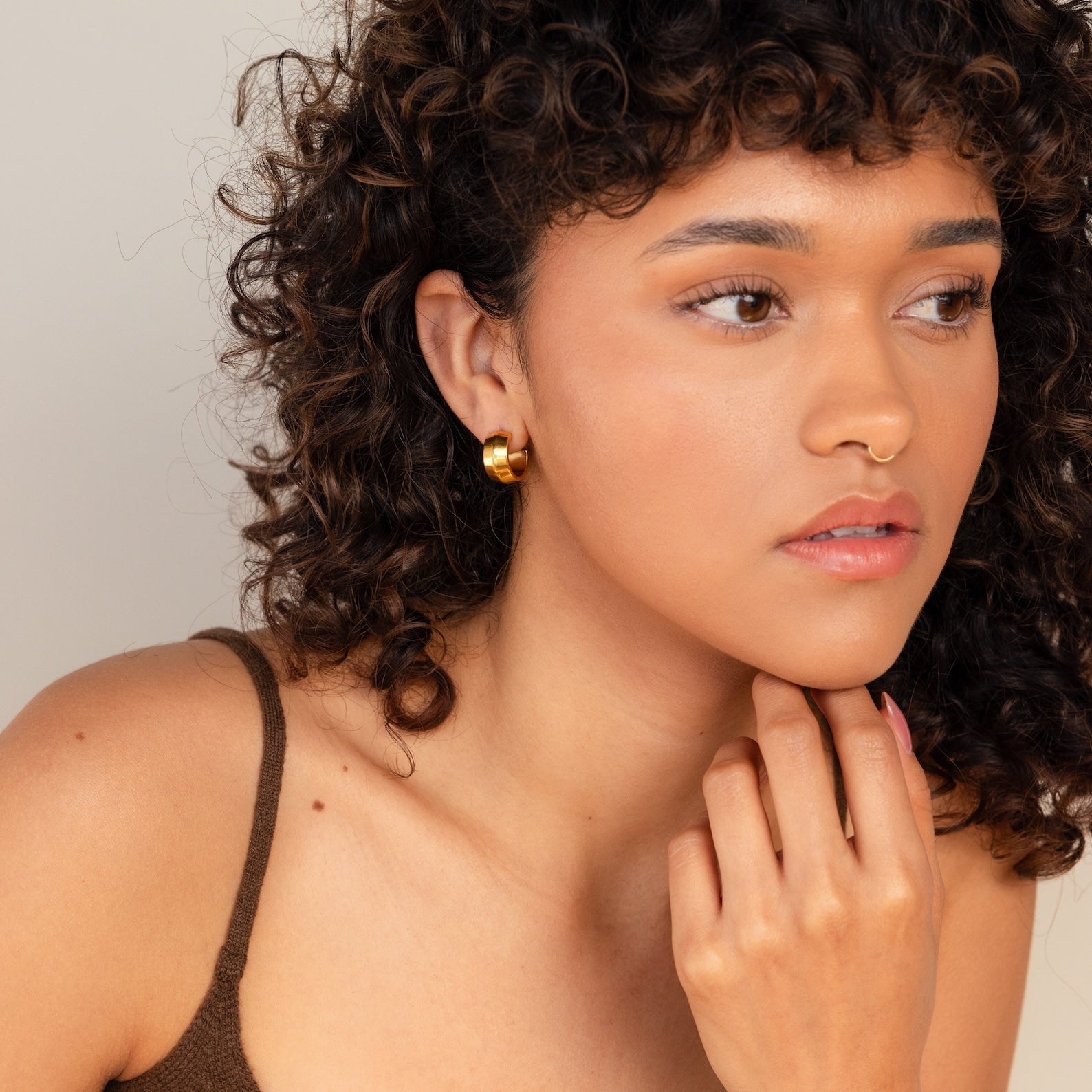 Woman with curly hair and statement jewelry, wearing a brown tank top and trendy Chunky Bold Hoops earrings, looking thoughtful.