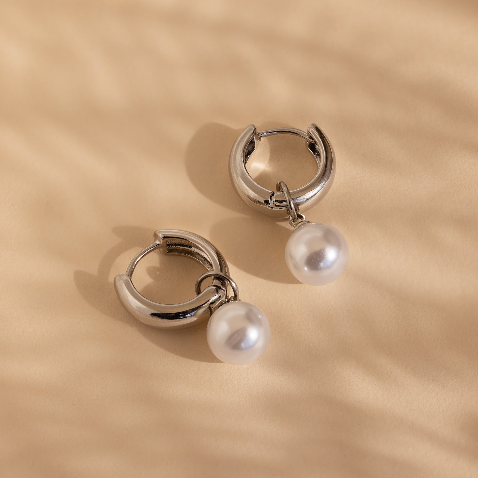 Bold Pearl Drop Earrings featuring silver hoops with dangling white pearls, perfect as statement pieces, showcased on a soft beige textured background.