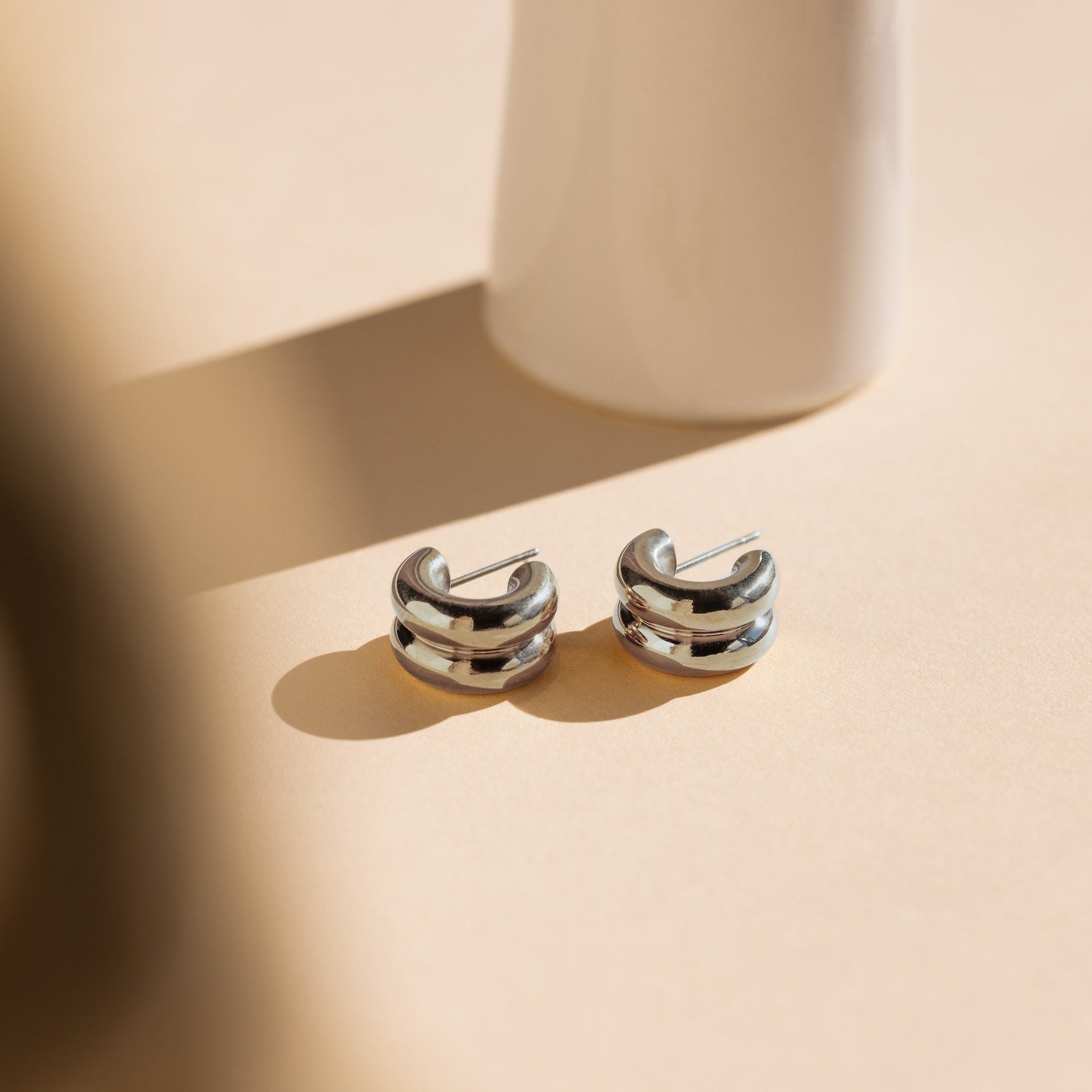 Chunky Double Hoops displayed on a beige surface with soft shadows and a white cylindrical backdrop—these statement earrings are perfect for lovers of minimalist jewelry.