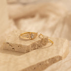 The Art Deco Ring Set features two delicate gold rings with clear gemstones, evoking vintage elegance, displayed on a textured stone surface against a soft beige background.