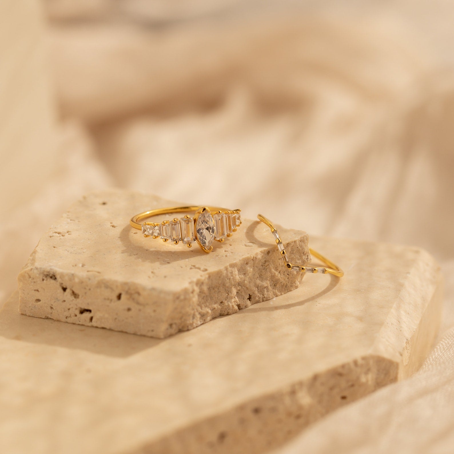 The Art Deco Ring Set features two delicate gold rings with clear gemstones, evoking vintage elegance, displayed on a textured stone surface against a soft beige background.