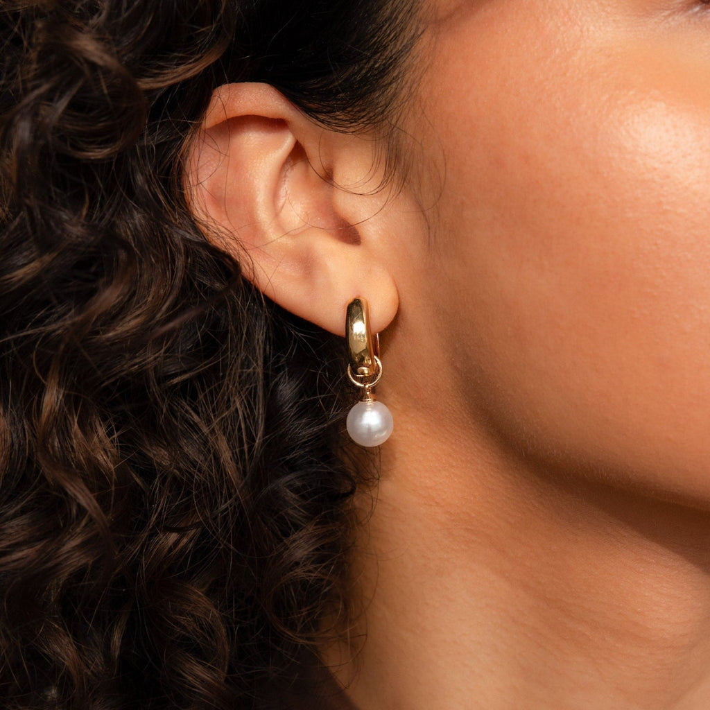 Close-up of a woman's ear wearing Bold Pearl Drop Earrings—gold hoops with a hanging pearl—her curls softly framing these statement earrings.